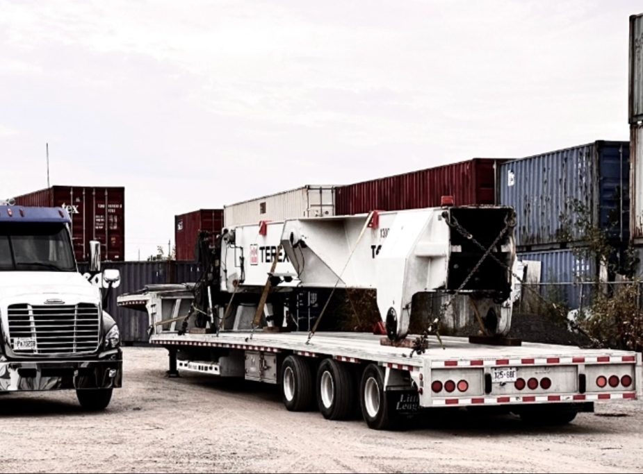 Step deck flatbed trailer hauling a heavy industrial machinery load at a shipping yard terminal.