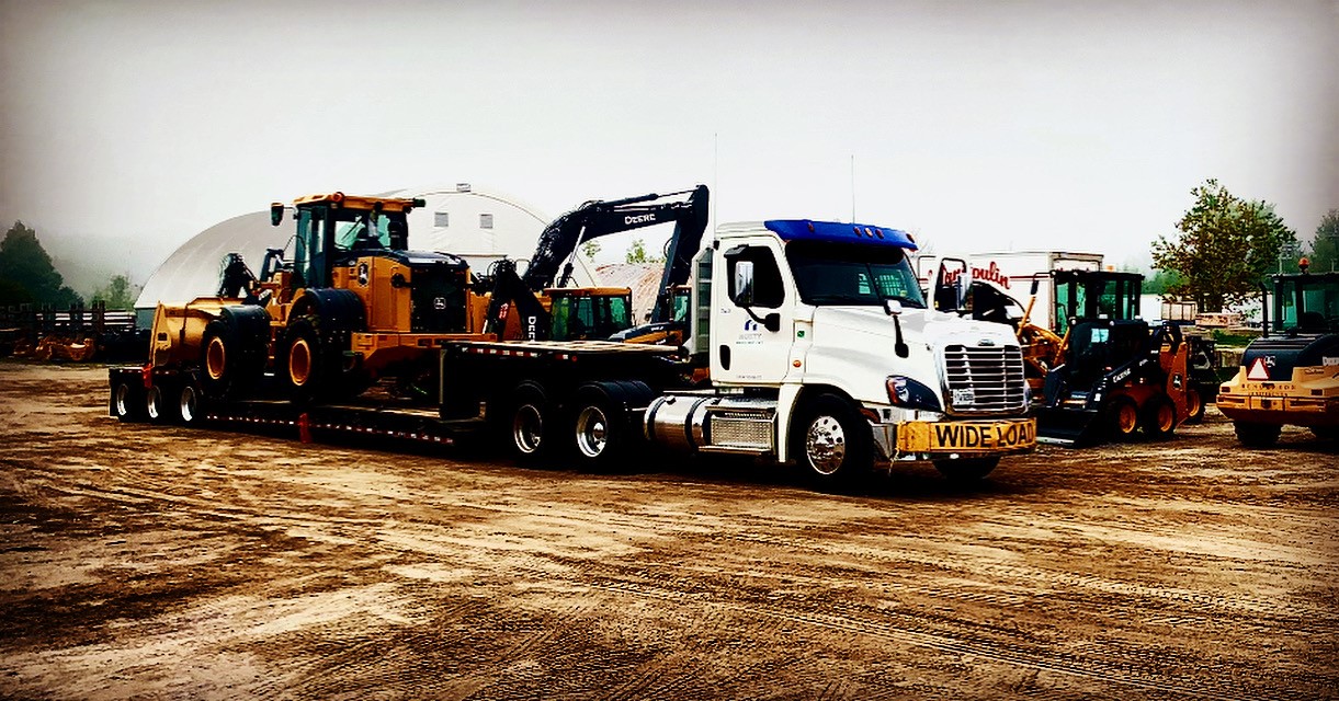A commercial semi truck hauling heavy construction machinery on a low-boy trailer in an industrial yard.